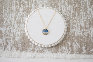 A delicate gold necklace with a small oval pendant featuring a scenic landscape of blue water and white sand, displayed on a white circular bead-edged stand over a textured beige and white fabric background.