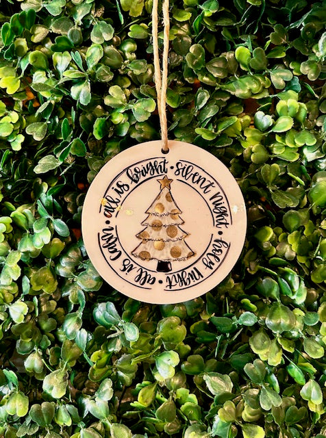Round wooden ornament with Christmas tree design and text, hanging on green foliage.