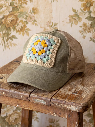 Green cap with a crochet flower patch on a wooden surface with floral wallpaper in the background