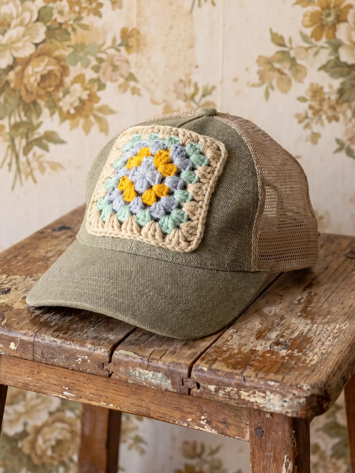 Green cap with a crochet flower patch on a wooden surface with floral wallpaper in the background