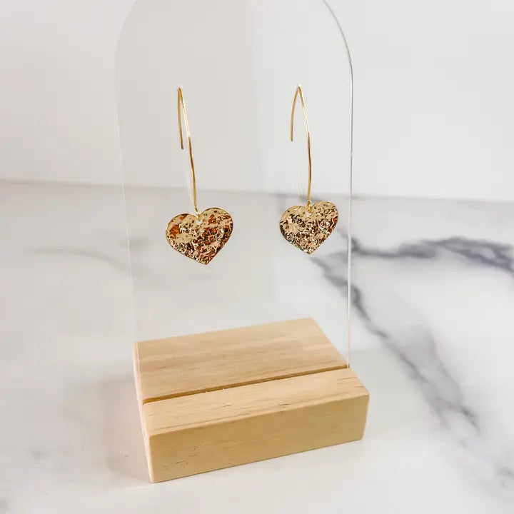 Heart-shaped gold earrings on a clear stand with a wooden block below on a marble surface.
