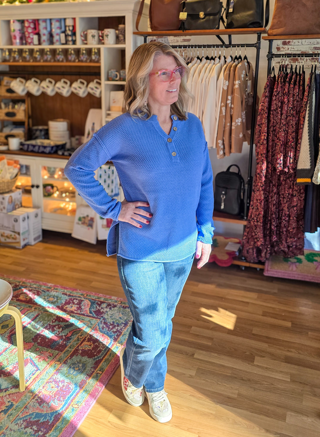 Woman in a blue shirt and jeans standing in a store with clothing racks and shelves in the background.