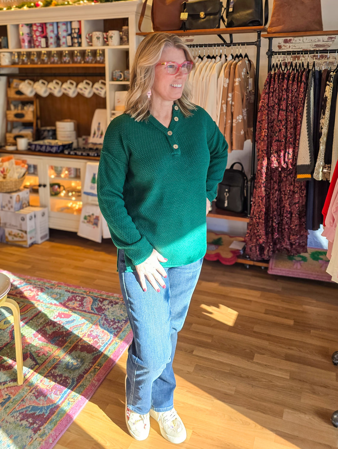 Woman in a green sweater and jeans standing in a store with clothing racks and displays.