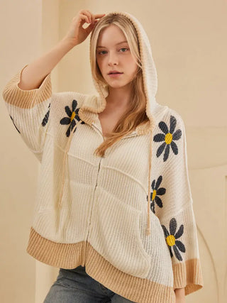A woman wearing a cream-colored ribbed knit zip-up hoodie with large black and yellow daisy flower appliqués on the sleeves and hood, paired with gray jeans.
