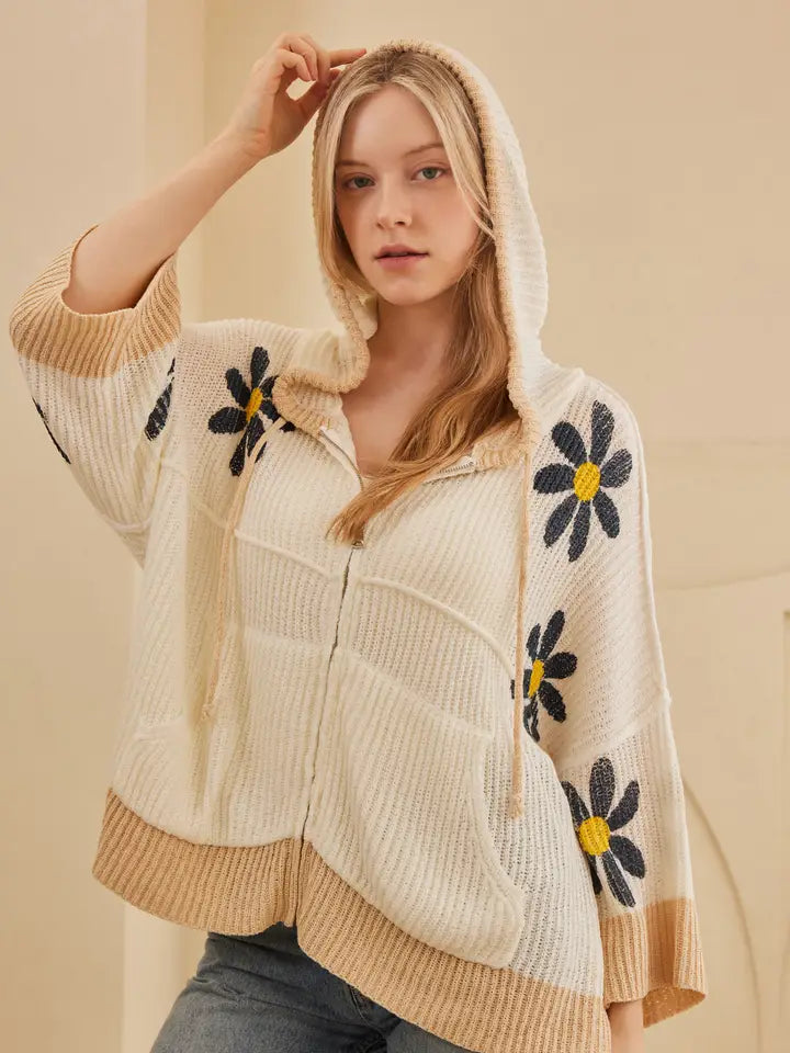 A woman wearing a cream-colored ribbed knit zip-up hoodie with large black and yellow daisy flower appliqués on the sleeves and hood, paired with gray jeans.