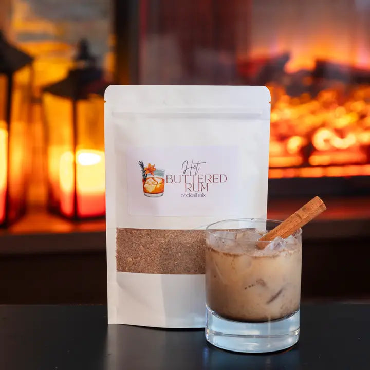 Buttered Rum cocktail mix package with a glass of cocktail in front of a fireplace.