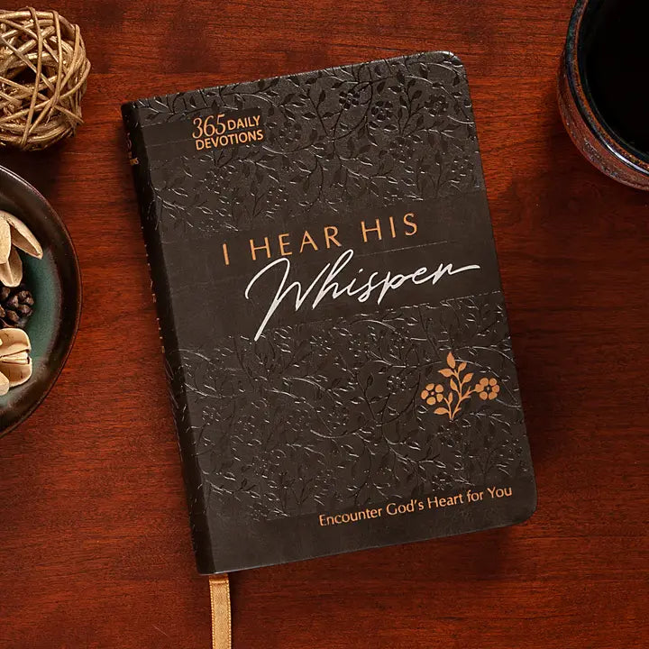 Dark leather devotional book titled 'I Hear His Whisper' on a wooden surface with a cup and decorative elements.