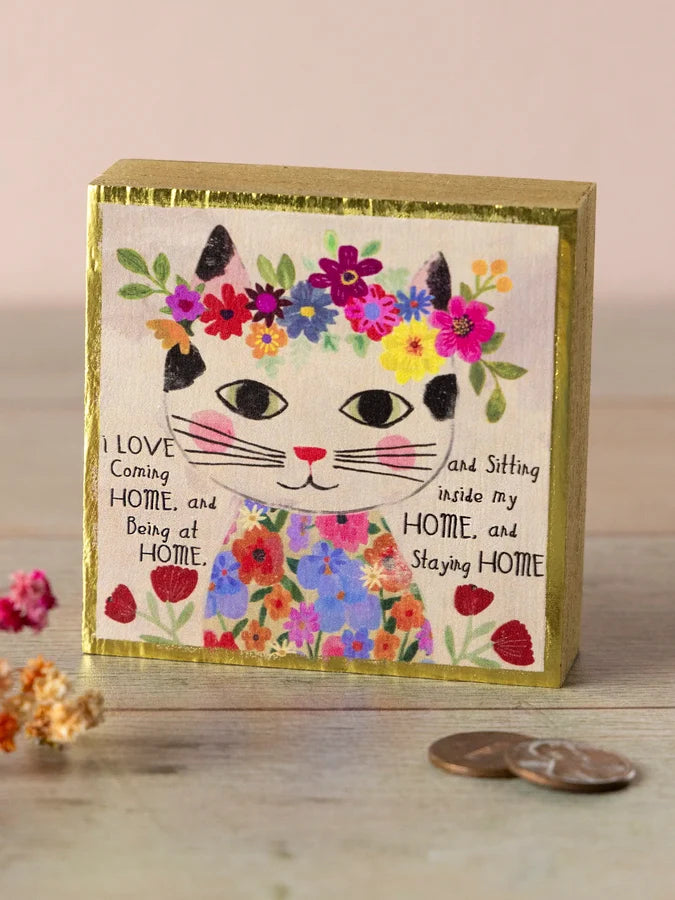 A square decorative block or plaque with a gold-toned border and a colorful hand-painted cat face surrounded by flowers, with the phrase &