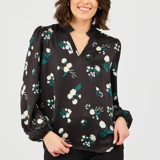 A woman wearing a black long-sleeve blouse with a white and green floral print, featuring a split V-neckline and gathered sleeves, photographed against a light gray studio background.