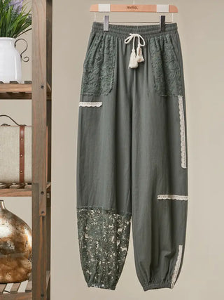 A pair of dark olive green patchwork jogger pants with lace details, hanging on a wooden hanger. The pants feature a drawstring waist, lace pocket trims, and lace hem details.