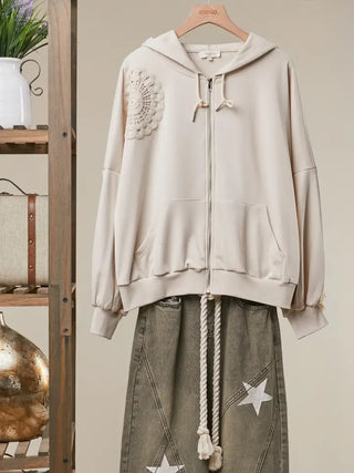 A beige zip-up hoodie with a crochet floral applique on the sleeve, displayed on a wooden hanger next to a gray denim skirt with white star patches.