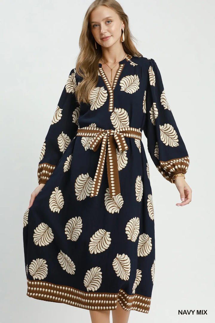 A woman modeling a navy blue long-sleeve midi dress with a beige palm leaf pattern and a brown belt with white dot accents, set against a plain light gray background.