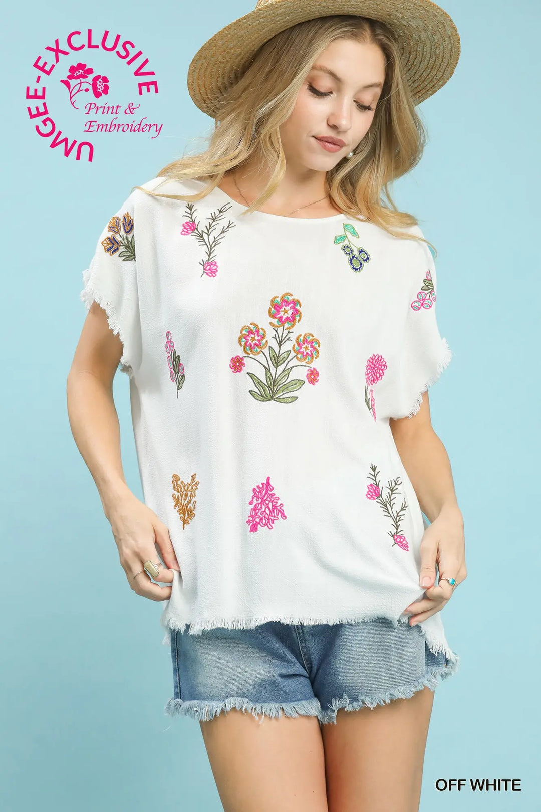 Woman wearing a white blouse with floral embroidery and denim shorts on a light blue background.