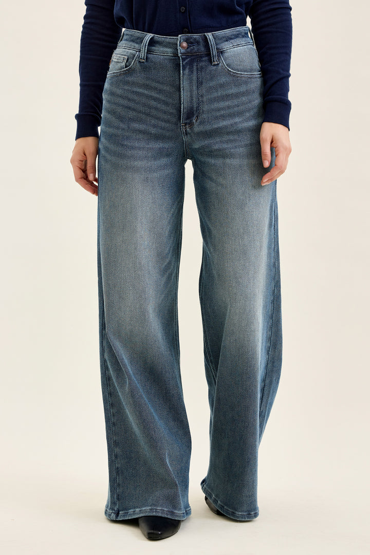 A person wearing high-waisted, wide-leg blue denim jeans with a faded wash, paired with a dark navy long-sleeve top and black shoes, against a plain light beige studio background.