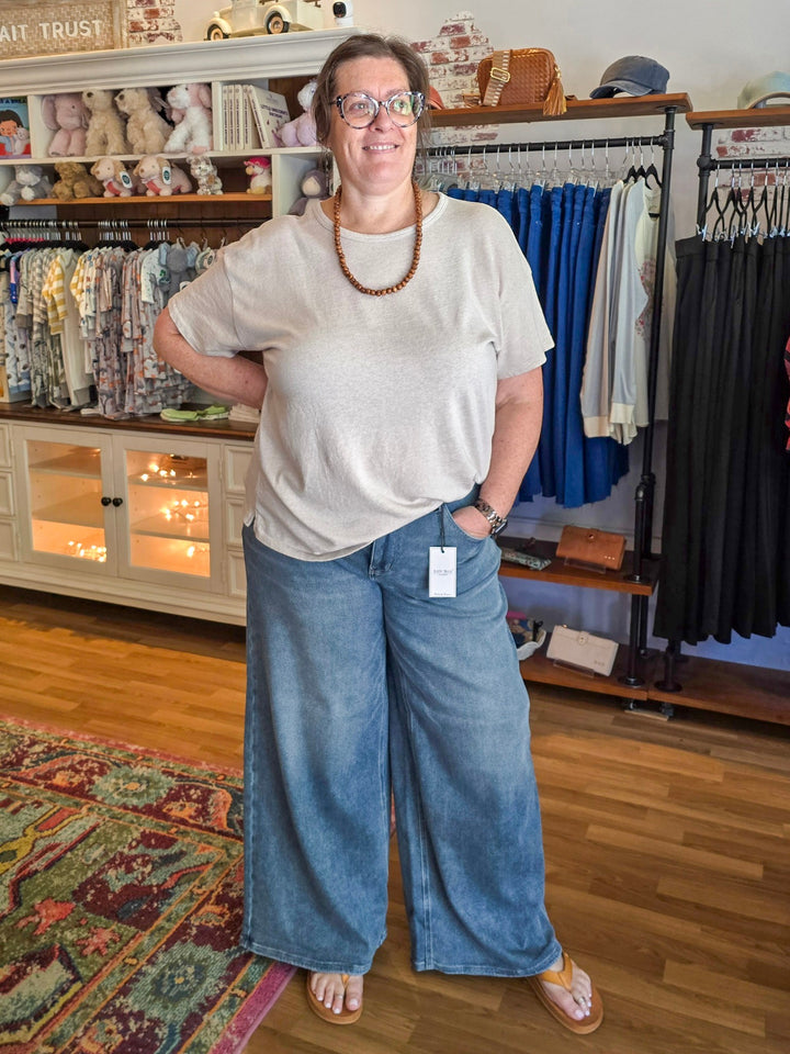 A woman wearing a light beige short-sleeve t-shirt and wide-leg blue denim jeans stands inside a boutique store. The background features clothing racks with various garments, shelves with stuffed animals, and a wooden floor.