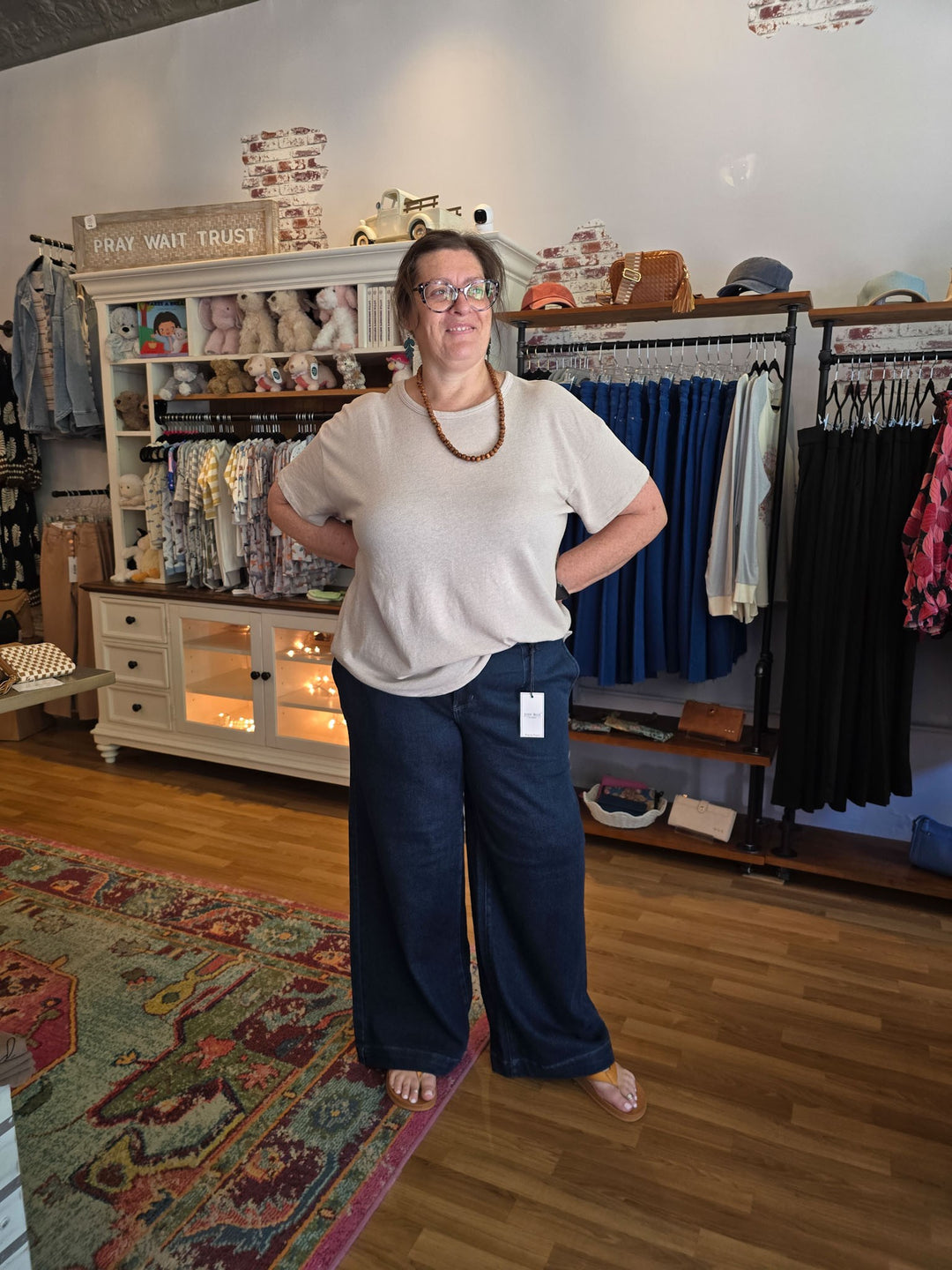 A woman wearing a cream short-sleeve top and navy wide-leg pants stands in a boutique store with clothing racks and a wooden floor behind her.