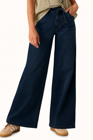 A person wearing dark navy wide-leg denim jeans with a drawstring waist, paired with a beige top and beige sneakers, shown from the waist down against a plain white background.