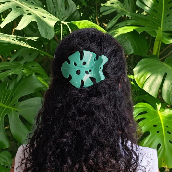 A mint green monstera leaf-shaped hair claw clip holding back long, curly black hair, shown from behind against a background of lush green tropical monstera leaves.