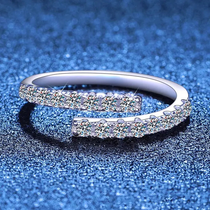 A silver-toned open band ring set with multiple round clear stones on both sides, photographed on a textured blue background.