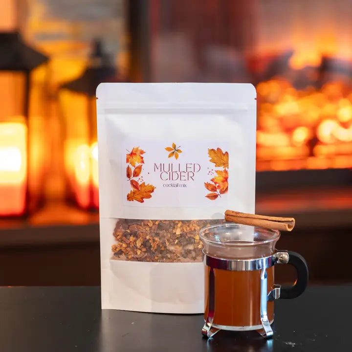 Packaged mulled cider with a mug of cider in front of a fireplace.