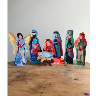 A set of hand-painted wooden nativity figurines arranged on a rustic wooden table against a plain off-white wall. The colorful figures include Mary, Joseph, baby Jesus in a manger, and three wise men.