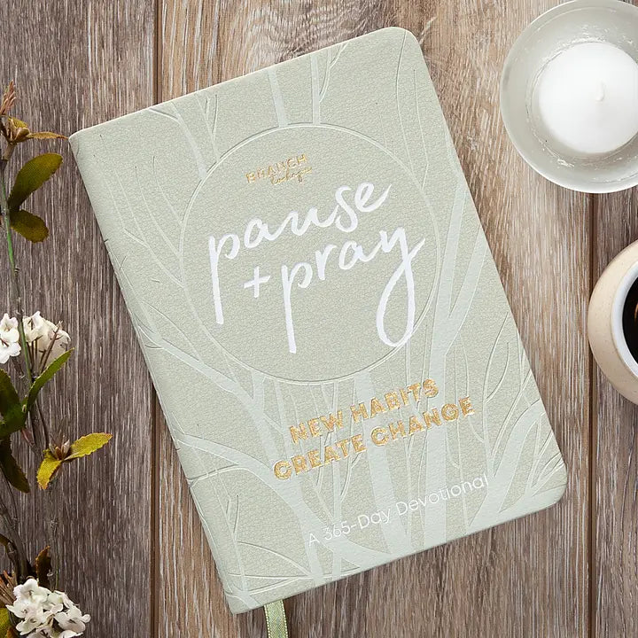 Journal with 'pause + pray' text on a wooden surface with flowers and a candle.