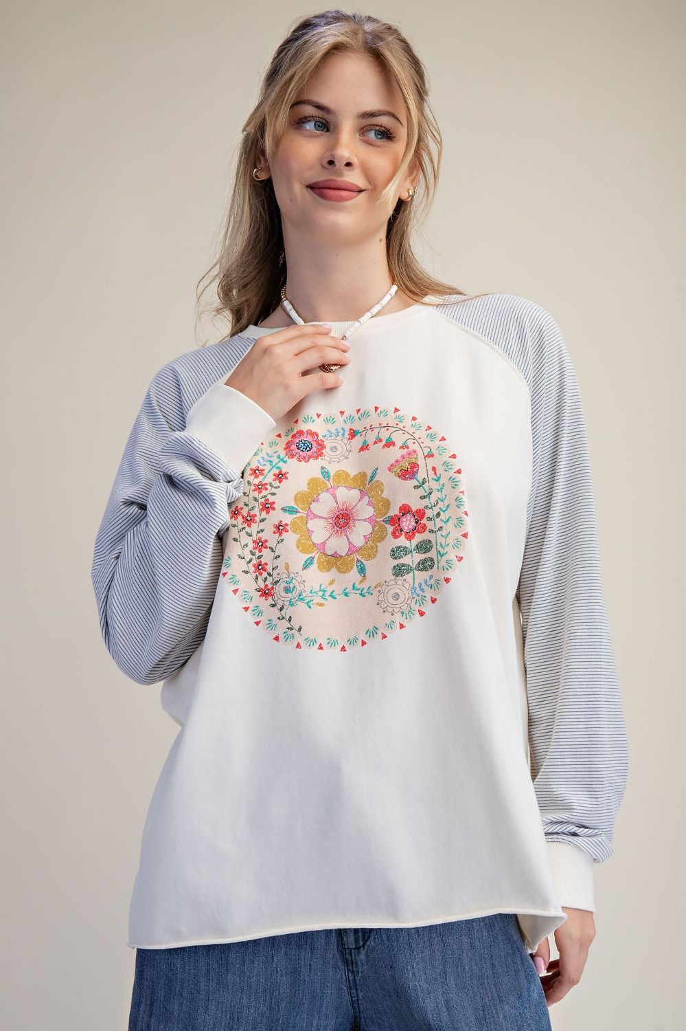 A woman wearing a white raglan sweatshirt with light blue striped sleeves and a colorful floral mandala graphic on the front, paired with blue jeans, against a solid beige background.