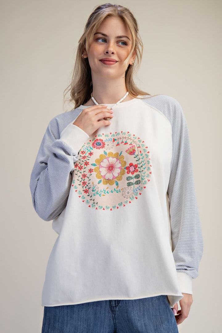 A woman wearing a white raglan sweatshirt with light blue striped sleeves and a colorful floral mandala graphic on the front, paired with blue jeans, against a solid beige background.