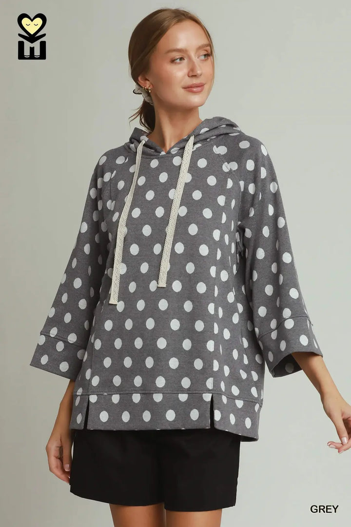 A woman wearing a grey hooded sweatshirt with a white polka dot pattern, featuring three-quarter sleeves and cream-colored drawstrings, paired with black shorts against a solid light grey background.