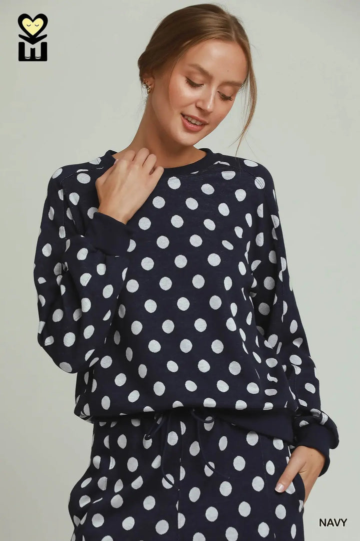 A woman wearing a navy blue long-sleeve top and matching pants with a white polka dot pattern, photographed against a light gray studio background. A small logo appears in the top-left corner, and the word &