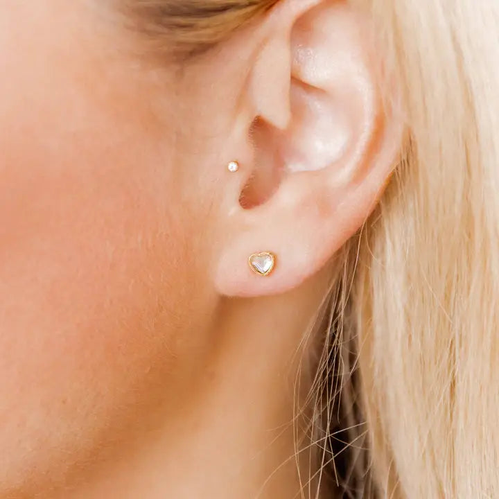 Close-up of an ear wearing a heart-shaped gold earring with blonde hair.