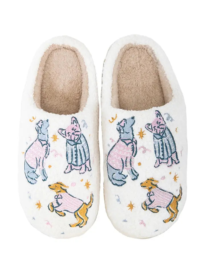 A pair of white plush slipper shoes decorated with embroidered dogs wearing pink and blue sweaters, set against a plain white background.