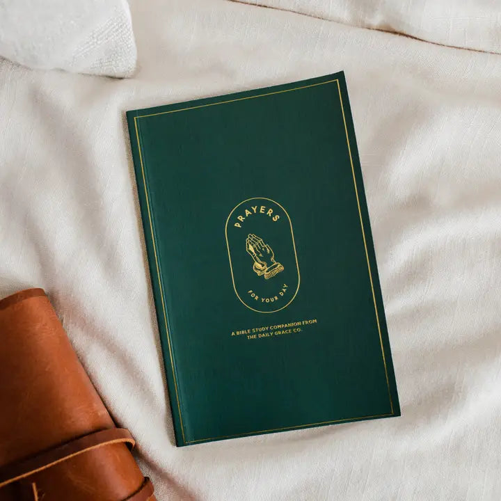 Green book titled 'Prayers of Your Day' on a white surface with a brown leather item beside it.