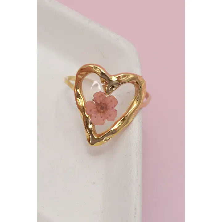 Gold heart-shaped ring with a pink flower on a white and pink background