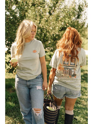 Two women walking outdoors in a grassy area, wearing matching light sage green graphic t-shirts and denim shorts. The woman on the left holds a green apple, and the woman on the right carries a black woven basket.