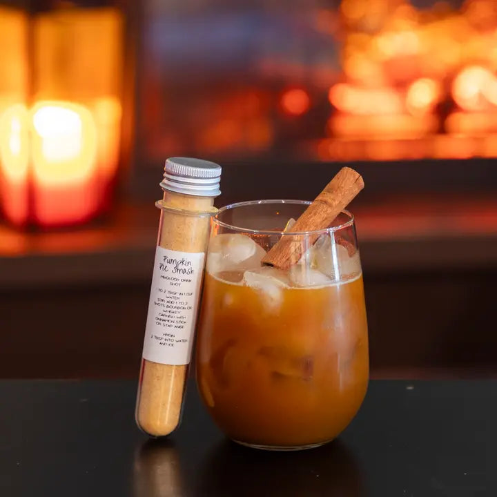 Glass of iced coffee with a cinnamon stick, bottle of pumpkin spice syrup, and blurred fireplace in the background.