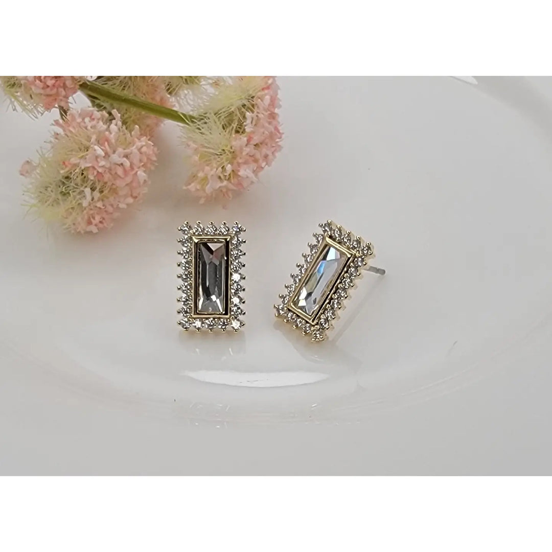 Pair of gold and crystal earrings on a white surface with pink flowers in the background