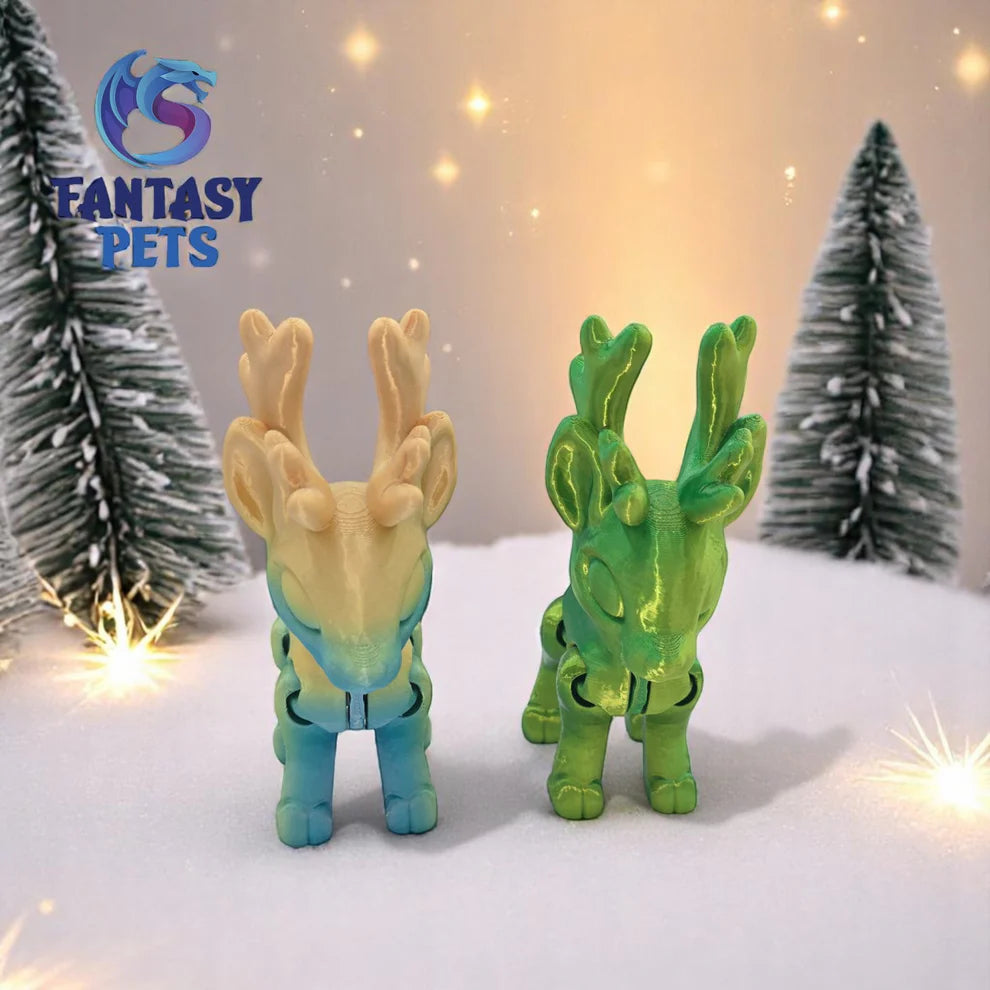Two toy deer figures, one beige and one green, on a snowy surface with Christmas trees and lights in the background. Fantasy Pets logo visible.