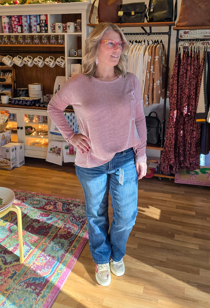Woman in a pink sweater and blue jeans standing in a store with clothing racks and shelves in the background.