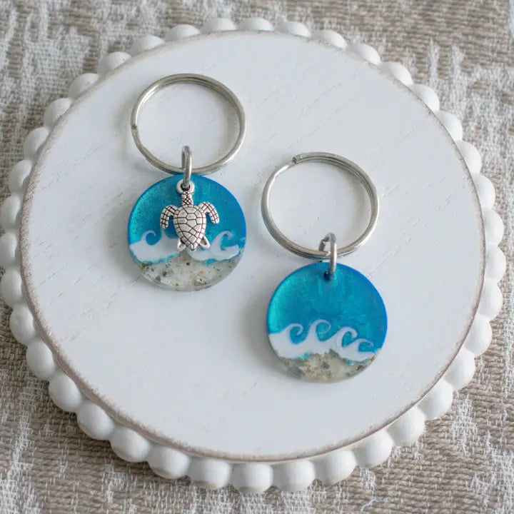 Two circular resin keychains with blue ocean wave designs and silver metal rings, displayed on a white scalloped wooden board over a textured beige fabric background.