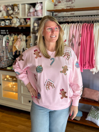 Blonde woman in pink sweatshirt with gingerbread men, gingerbread house, Christmas ornaments scattered throughout. 
