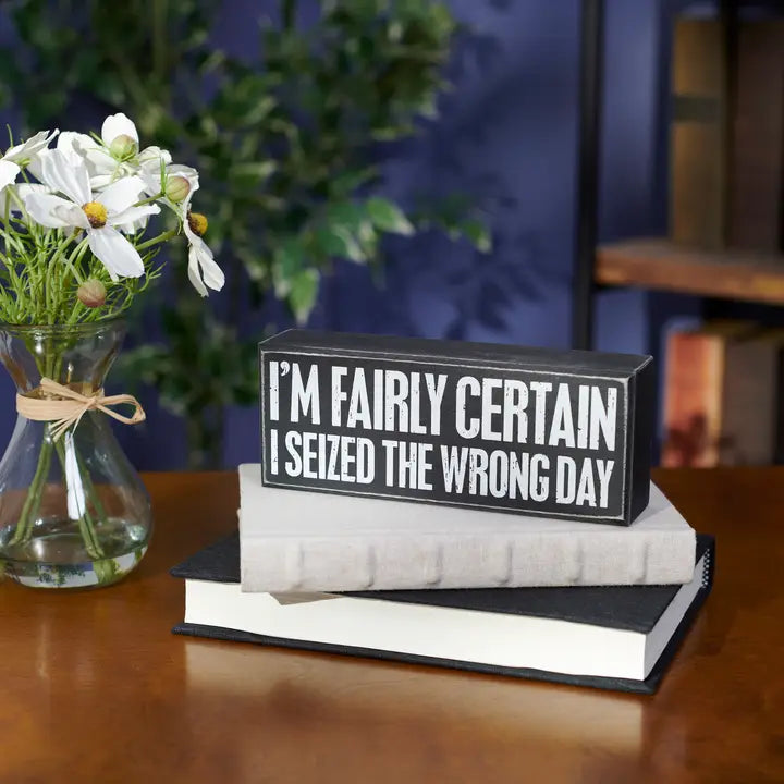 Decorative sign on books with text 'I'm fairly certain I seized the wrong day' next to a vase with flowers.