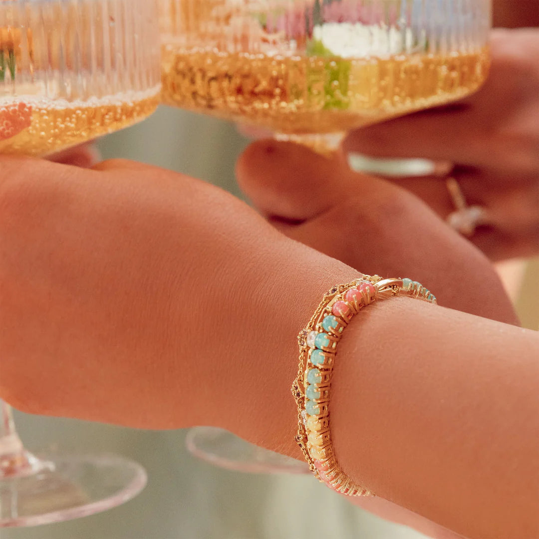 Close-up of a hand wearing a gold bracelet with colorful stones, holding a glass with a floral arrangement.