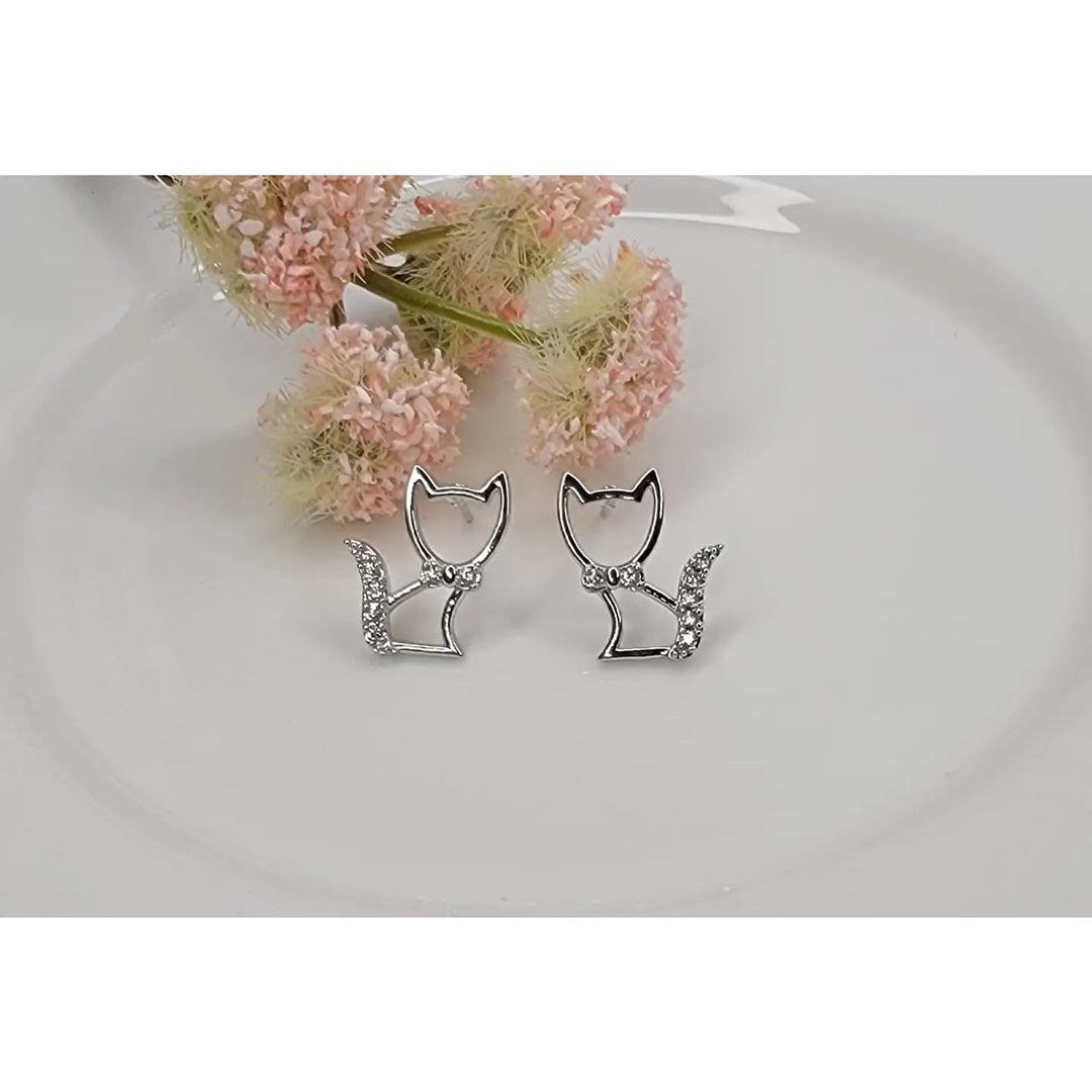 Cat-shaped earrings with rhinestones on a white surface with pink flowers in the background