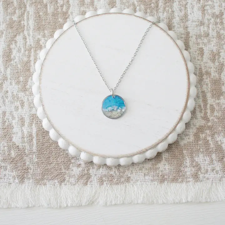 A silver necklace with a round pendant featuring a blue and white landscape design, displayed on a white scalloped circular stand over a textured beige fabric background.