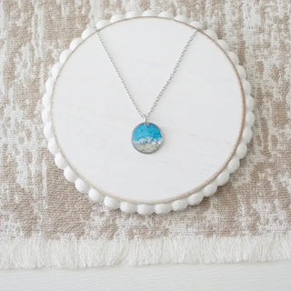 A silver necklace with a round pendant featuring a blue and white landscape design, displayed on a white scalloped circular stand over a textured beige fabric background.