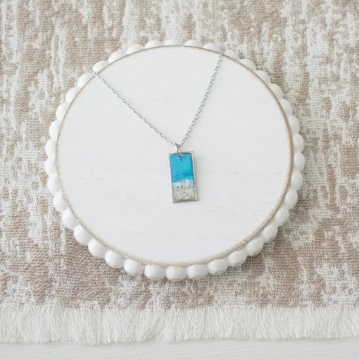 A silver necklace with a rectangular pendant featuring a blue sky and white clouds design, displayed on a round white wooden stand over a textured beige fabric background.