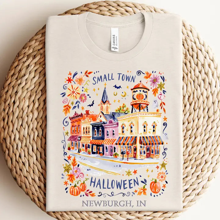 A folded cream-colored t-shirt with a colorful watercolor-style graphic of a small town Halloween scene, displayed on a round woven straw mat. The graphic includes the text &