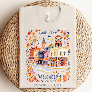 A folded cream-colored t-shirt with a colorful watercolor-style graphic of a small town Halloween scene, displayed on a round woven straw mat. The graphic includes the text 'SMALL TOWN HALLOWEEN NEWBURGH, IN'.