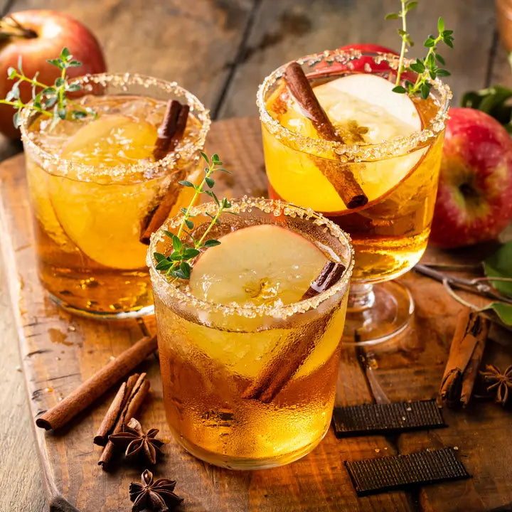 Three glasses of apple cider with cinnamon sticks and apples on a wooden surface.
